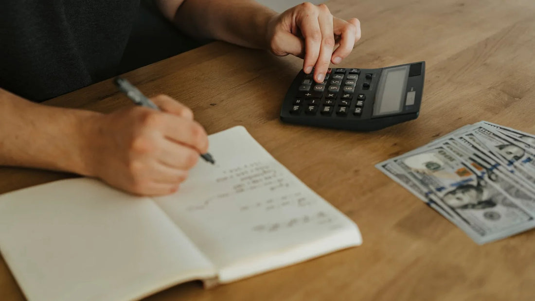 Close-up of hands using a calculator and writing in a notebook next to cash on a wooden table