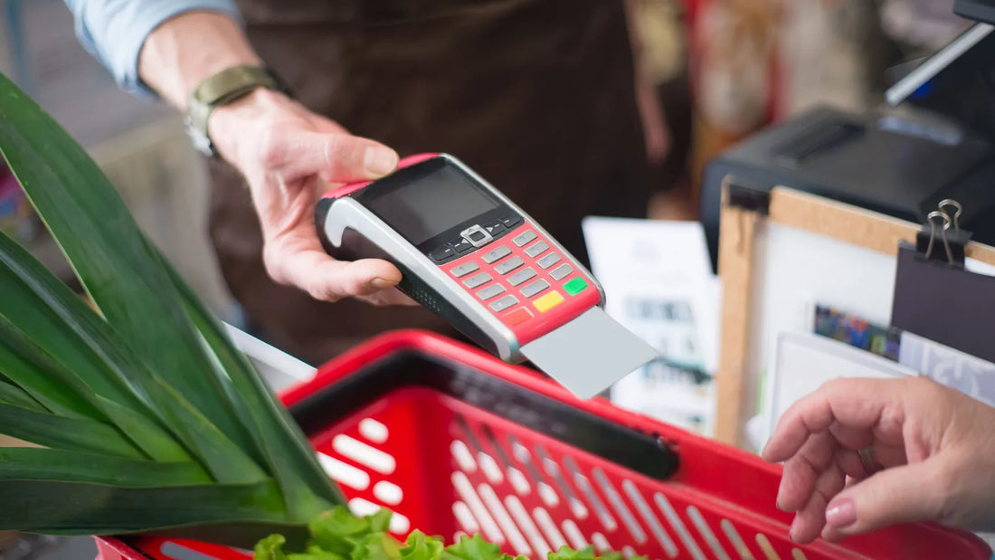 Shopper paying for groceries with a plain card at a checkout terminal next to a basket of vegetables