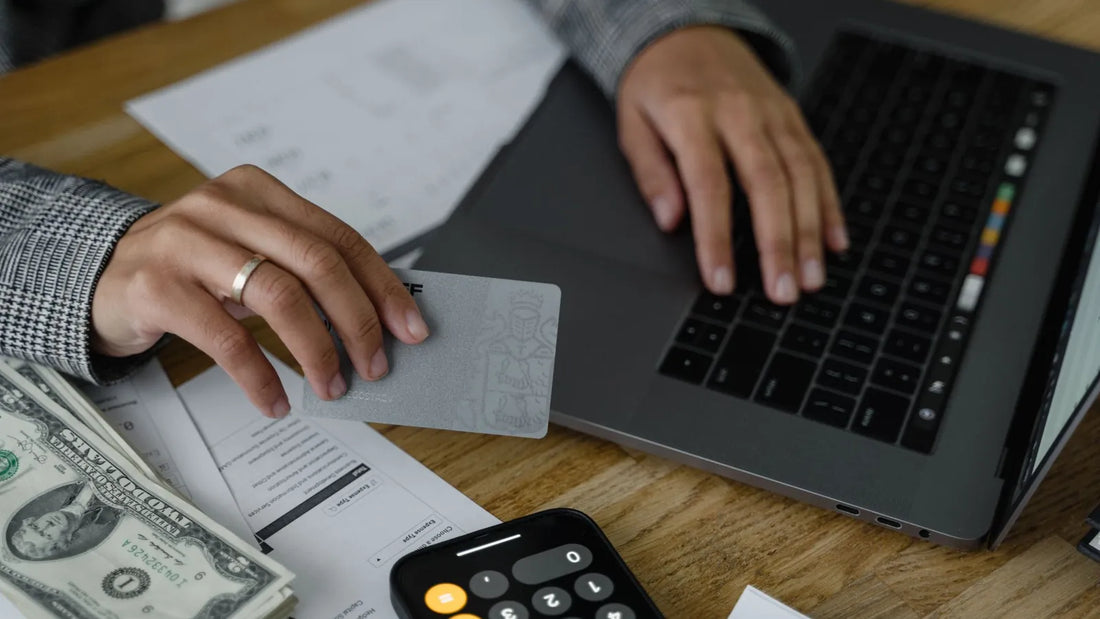 Hands holding a payment card over a laptop, with cash and a phone calculator on the desk