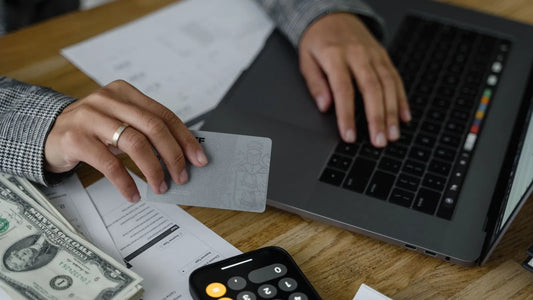 Hands holding a payment card over a laptop, with cash and a phone calculator on the desk