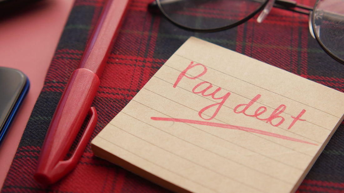 Handwritten note reading Pay debt next to a pen and glasses, representing a debt payoff plan