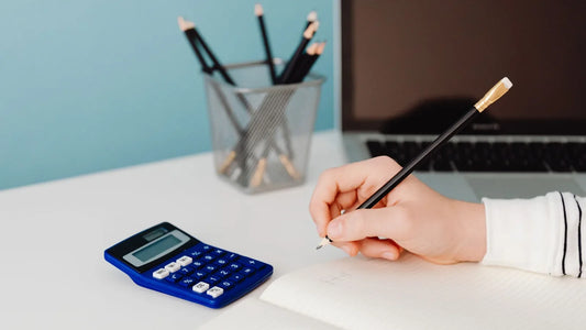 Hand writing in a notebook at a desk with a calculator and laptop