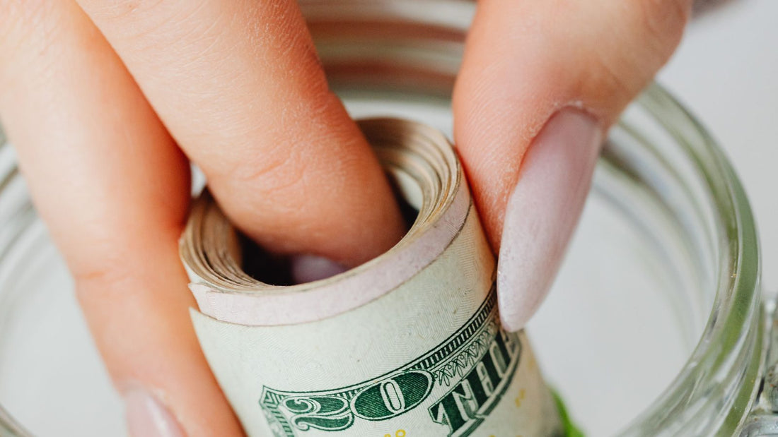 Close-up of a hand placing a rolled twenty dollar bill into a glass savings jar for an emergency fund