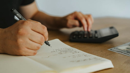 Person handwriting expense calculations in a notebook next to a calculator and cash