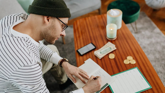 Person writing in a small notebook at a coffee table with cash and a phone nearby, doing a weekly money check-in