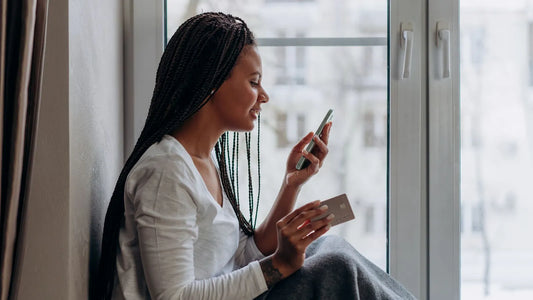 Woman sitting by a window holding a credit card and looking at her phone