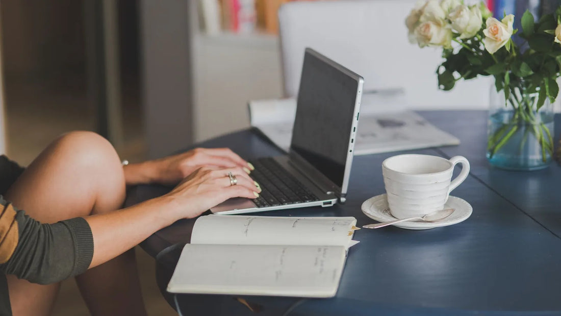 Person typing on a laptop next to an open notebook and coffee cup at a home desk, planning a budget with multiple income sources