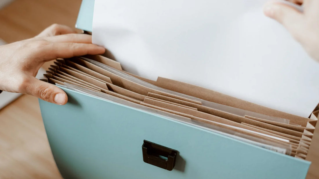 Hands pulling a sheet of paper from a labeled accordion file folder on a wooden table