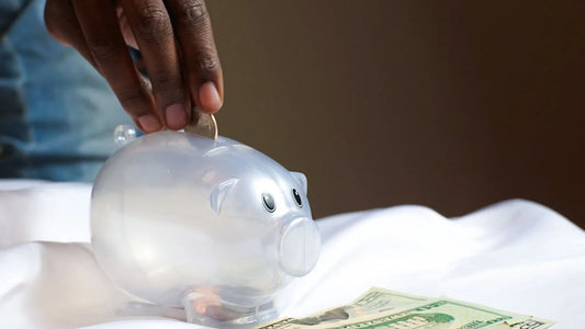 Hand placing a coin into a clear piggy bank next to dollar bills, representing rebuilding an emergency fund