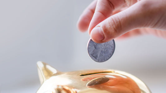 Hand dropping a coin into a gold piggy bank, representing saving money on a tight budget