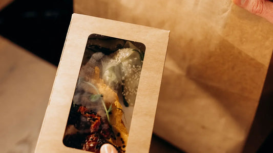 Hands holding a cardboard takeout container next to a paper delivery bag