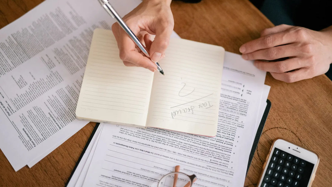 Hands writing in a notebook surrounded by tax paperwork and a phone